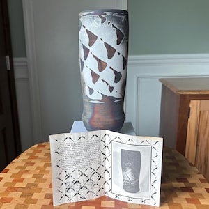 May include: A cylindrical ceramic vase with a matte finish, featuring a pattern of dark brown and black triangular shapes against a white background. The base is a solid, dark metallic brown. An informational card with text is placed in front.