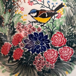 May include: A ceramic plate with a colorful floral and bird design. The plate features a blue, yellow, and brown bird perched on a branch, surrounded by red roses, a blue flower, and green foliage. The background is white.