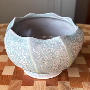 May include: A ceramic bowl with a scalloped rim and a rounded body. The exterior features a speckled light green and white glaze with subtle yellow vertical accents. The interior is smooth and light gray, and the bowl sits on a circular base.