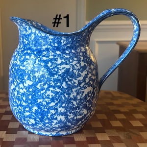 May include: Blue and white speckled ceramic pitcher with a curved handle. The pitcher has a wide mouth and a dark blue rim. The background is blurred, but the pitcher is sitting on a wooden surface.