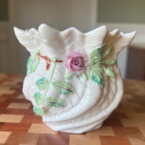 May include: Cream-colored ceramic vase with a seashell design. The vase features a pink rose and green leaves with blue accents. The vase has a wavy rim and a textured, swirled body, perfect for home decor.