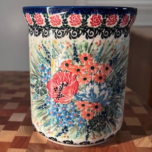 May include: A ceramic utensil holder with a floral design. The holder features a blue rim and is decorated with red poppies, orange and blue flowers, and green foliage. The base has a black and white pattern.