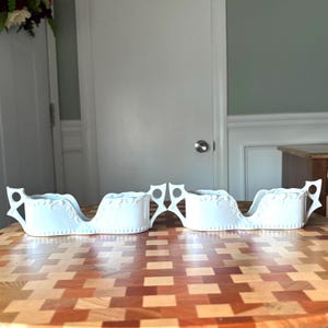 May include: Two matching white ceramic serving dishes. Each dish has a sleigh-like shape with decorative floral patterns and handles. The dishes are placed on a wooden surface with a geometric pattern.