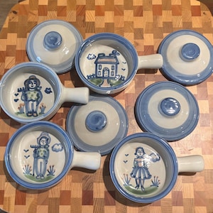 May include: A collection of three ceramic bowls with handles and three matching lids. Each bowl features a hand-painted blue and white design, including illustrations of a house, a person with a rake, and a person in a dress, all with blue rims.