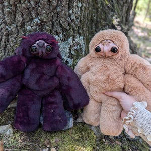 May include: Two plush creatures with soft faux fur, one in deep purple and one in light tan. Each features a sculpted, smooth face with large, dark, glossy eyes and a small beak-like nose. The purple creature has thin red antennae on its head.