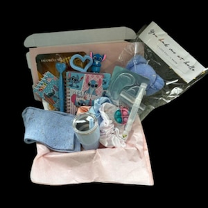 May include: A gift box filled with various items, including a blue tumbler, notebooks, a pen, a heart-shaped item, a Stitch figurine, and a small pop-it toy. The box is lined with pink paper and includes a clear bag with text.