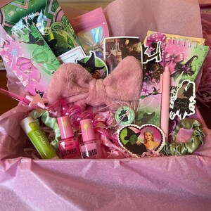 Puede incluir: Una caja de regalo rosa llena de varios artículos. Incluye un lazo rosa, esmalte de uñas, bálsamo labial, pegatinas, un bolígrafo y un pequeño cuaderno. La caja está forrada con papel de seda rosa, y los artículos tienen un tema de brujas.