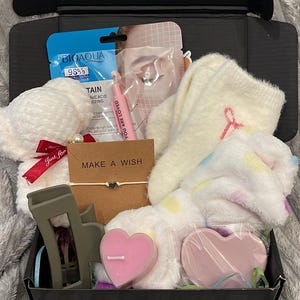 May include: A black gift box containing self-care products. Items include a white teddy bear, face mask, socks, a pink heart-shaped candle, a bracelet with "MAKE A WISH", and a hair clip. The box rests on a grey blanket.