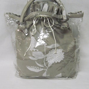 Può includere: Una borsa beige con coulisse con un motivo floreale bianco, racchiusa in un sacchetto di plastica trasparente.