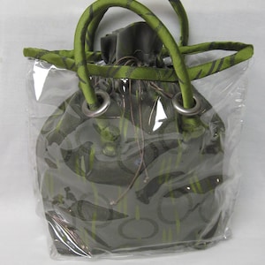 Può includere: Una borsa a cordoncino verde e nera con una copertura in plastica trasparente. La borsa ha due anelli metallici nella parte superiore.