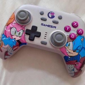 Puede incluir: Un mando Gamesir blanco con ilustraciones de Sonic the Hedgehog y Amy Rose. El mando tiene joysticks grises, una cruceta negra y botones rosas, morados y grises. El logotipo de Gamesir es visible.