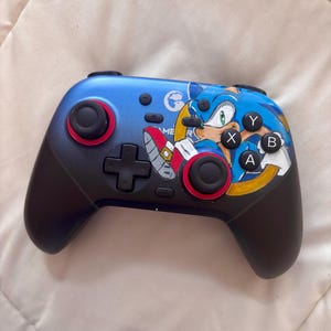 Puede incluir: Un mando de videojuegos azul y negro con un dise&ntilde;o de Sonic el Erizo. El mando tiene detalles rojos alrededor de los joysticks y botones. Los botones est&aacute;n etiquetados con las letras X, Y, A y B. Tambi&eacute;n presenta una cruceta y el texto "GAME ME R".