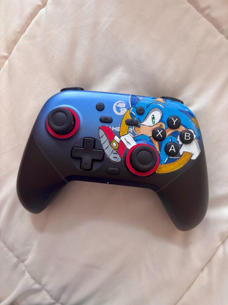 Puede incluir: Un mando de videojuegos azul y negro con un dise&ntilde;o de Sonic el Erizo. El mando tiene detalles rojos alrededor de los joysticks y botones. Los botones est&aacute;n etiquetados con las letras X, Y, A y B. Tambi&eacute;n presenta una cruceta y el texto "GAME ME R".