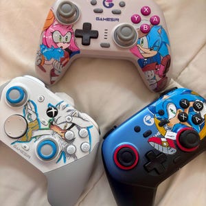 Puede incluir: Tres mandos de videojuegos con ilustraciones de personajes. Uno es blanco con Amy Rose, otro es blanco con Silver the Hedgehog, y el tercero es azul con Sonic the Hedgehog. Cada mando tiene un logotipo de Gamesir y varios botones.