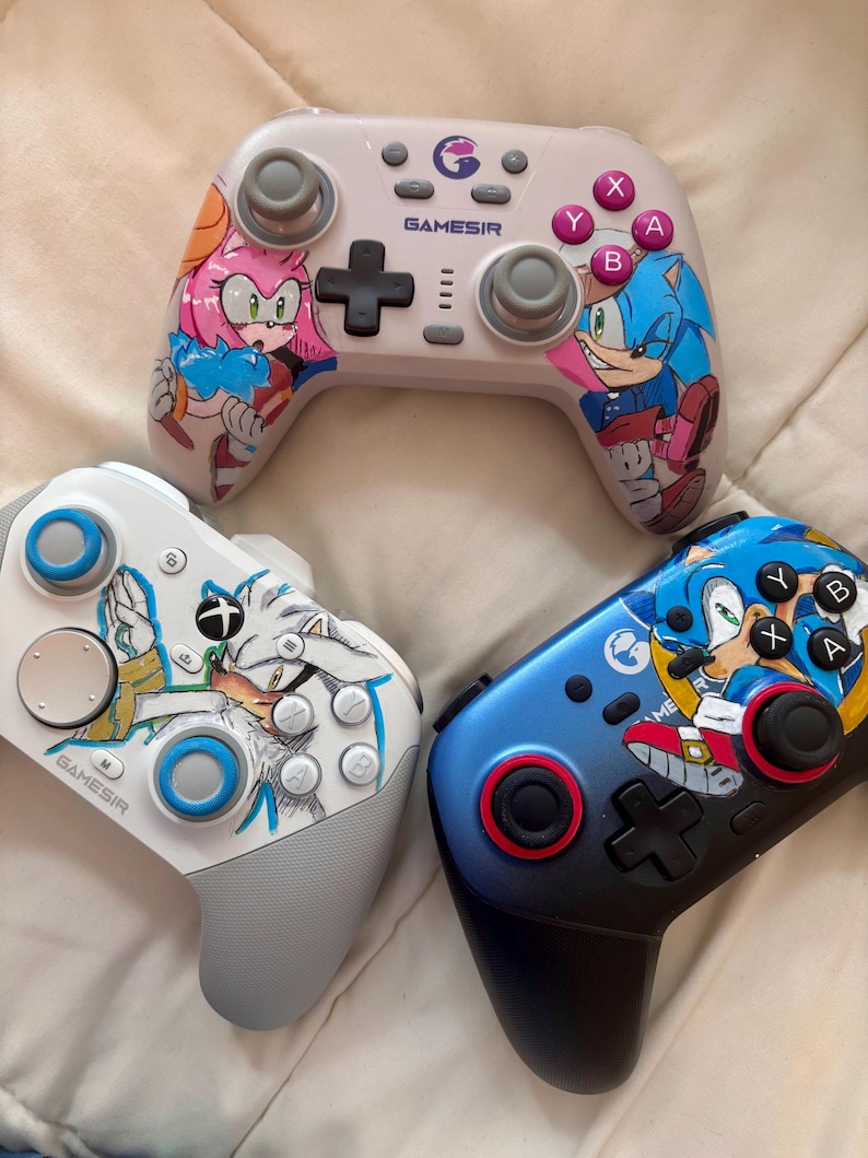 Puede incluir: Tres mandos de videojuegos con ilustraciones de personajes. Uno es blanco con Amy Rose, otro es blanco con Silver the Hedgehog, y el tercero es azul con Sonic the Hedgehog. Cada mando tiene un logotipo de Gamesir y varios botones.