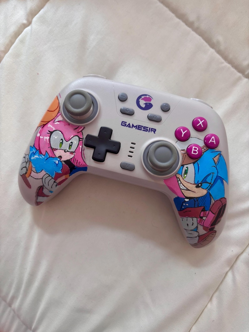 Puede incluir: Un mando Gamesir blanco con ilustraciones de Sonic the Hedgehog y Amy Rose. El mando tiene joysticks grises, una cruceta negra y botones rosas, morados y grises. El logotipo de Gamesir es visible.