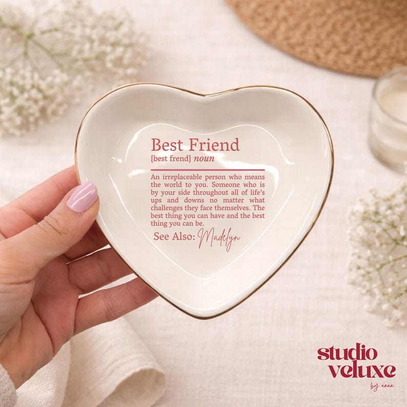 K&ouml;nnte beinhalten: Eine herzf&ouml;rmige Keramikschale mit einem goldfarbenen Rand. In der Mitte befindet sich ein aufgedruckter Text, der den Begriff Best Friend definiert, gefolgt von dem Namen Madelyn in Schreibschrift. Die Schale ist cremewei&szlig; mit dunkelrosa Schrift.