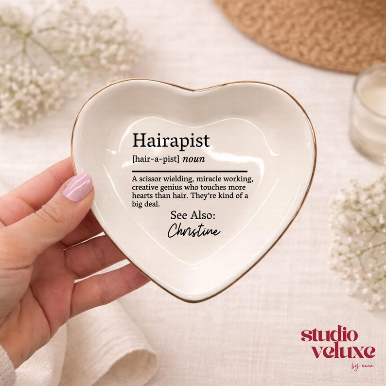 May include: A heart-shaped white ceramic trinket dish with a gold-colored rim. The center features black text reading: Hairapist [hair-a-pist] noun. A scissor wielding, miracle working, creative genius who touches more hearts than hair. See Also: Christine.