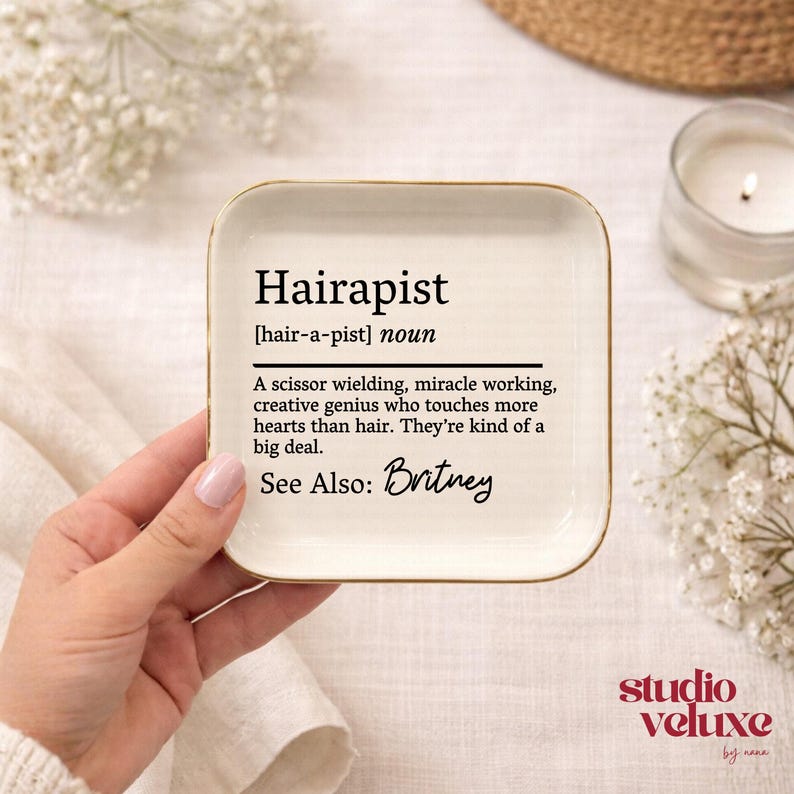 May include: A square white ceramic trinket dish with rounded corners and a gold-toned rim. The center features black printed text defining the word Hairapist as a scissor wielding, miracle working, creative genius, with the added text See Also: Britney.