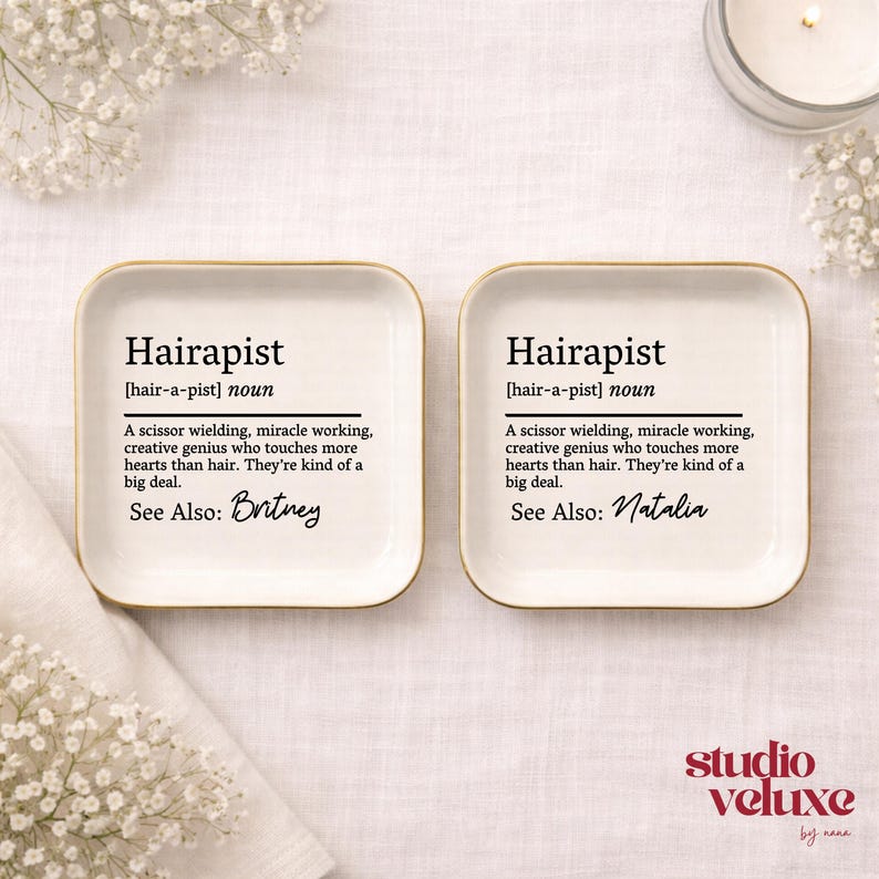 May include: Two square white ceramic trinket dishes with gold-toned rims. Each dish features black printed text defining the word Hairapist as a scissor wielding, miracle working, creative genius. One dish is personalized with Britney, the other Natalia.