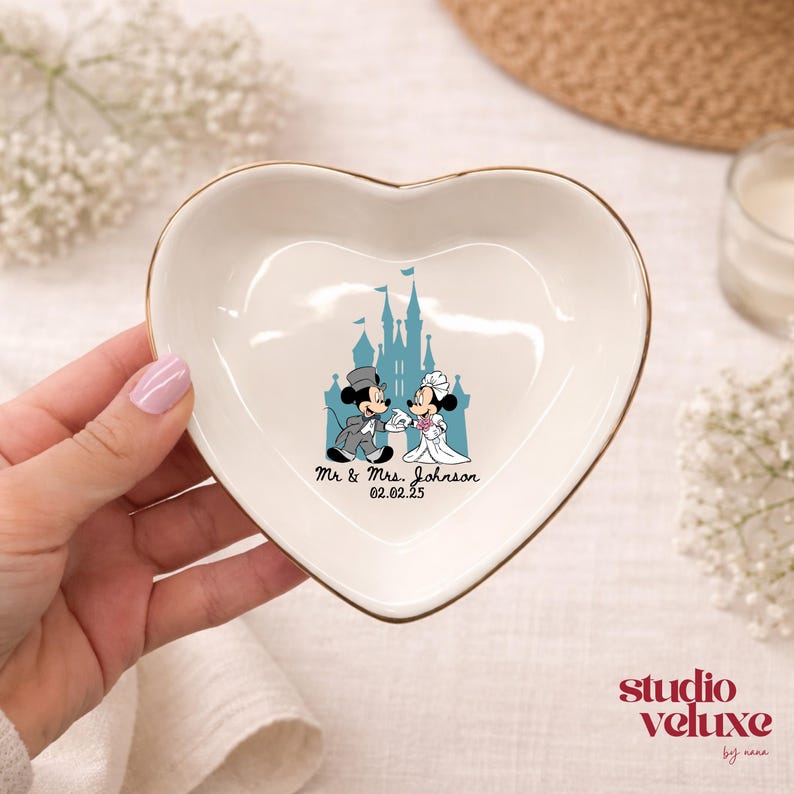 May include: Heart-shaped ceramic trinket dish with a gold rim. The dish features a cartoon image of Mickey and Minnie Mouse in wedding attire in front of a castle. The text "Mr & Mrs. Johnson 02.02.25" is printed below the image.