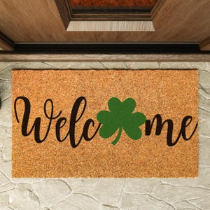 May include: A rectangular doormat made of natural coir fiber. The mat features the word "Welcome" in a black, cursive font, with a green shamrock replacing the "o" in the word. The mat is placed in front of a wooden door.