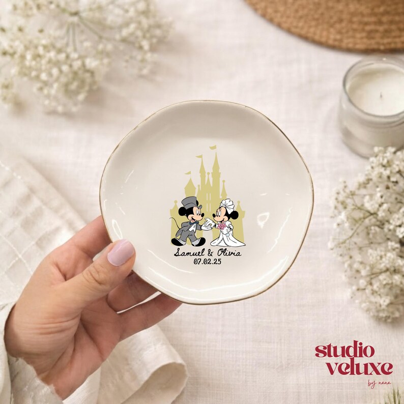 May include: A white ceramic ring dish with a gold rim. The dish features a cartoon image of Mickey and Minnie Mouse in wedding attire, with a castle backdrop. The names "Samuel & Olivia" and the date "07.02.25" are printed below.