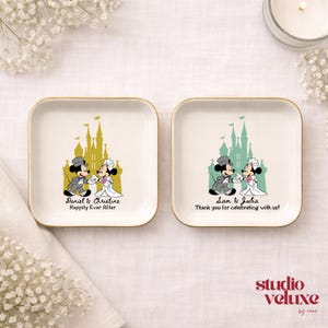 May include: Two square, white ceramic dishes with gold rims. Each features a cartoon couple in wedding attire in front of a castle. One says "Daniel & Christine Happily Ever After," the other says "Sam & Julia Thank you for celebrating with us!"
