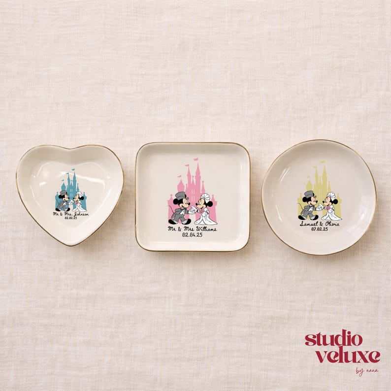 May include: Three white ceramic dishes with gold rims. One is heart-shaped, one square, and one round. Each features a Disney-themed wedding design with Mickey and Minnie Mouse, a castle, and names with a date.
