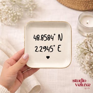 May include: A square, off-white ceramic trinket dish with gold trim. The dish features black text: "48.8584° N 2.2945° E" and a heart symbol. The Studio Veluxe logo is in the bottom right corner.