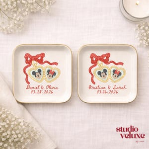 May include: Two square, off-white ceramic dishes with gold rims. Each dish features a red bow, Mickey and Minnie Mouse in heart-shaped frames, and names with a date. The dishes are on a white surface.