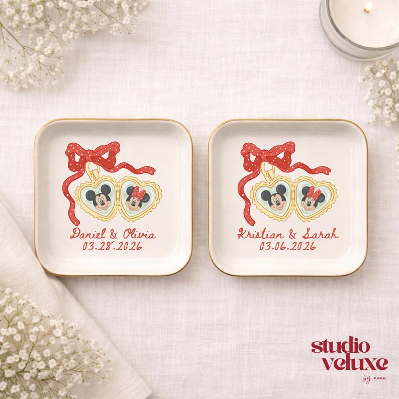 May include: Two square, off-white ceramic dishes with gold rims. Each dish features a red bow, Mickey and Minnie Mouse in heart-shaped frames, and names with a date. The dishes are on a white surface.