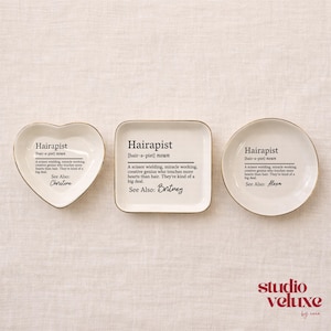 May include: Three white ceramic trinket dishes in heart, square, and round shapes with gold rims. Each dish features black printed text defining the word Hairapist, followed by a specific name: Christine, Britney, or Alexa, in script font.