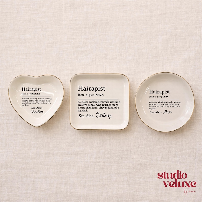 May include: Three white ceramic trinket dishes in heart, square, and round shapes with gold rims. Each dish features black printed text defining the word Hairapist, followed by a specific name: Christine, Britney, or Alexa, in script font.