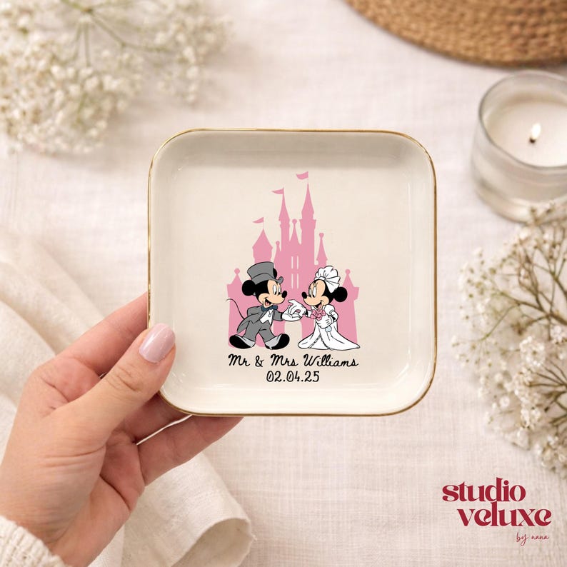May include: A square, off-white ceramic trinket dish with a gold rim. The dish features a pink castle graphic and a cartoon couple. Text on the dish reads "Mr. & Mrs. Williams 02.04.25".