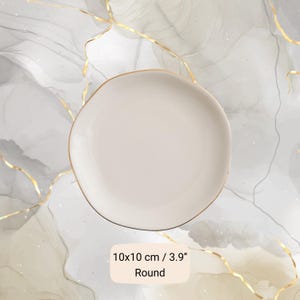 May include: A round, off-white plate with a gold rim. The plate measures 3.9 inches in diameter and has the word "Round" printed on the bottom. The plate is on a marble-like background.