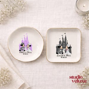 May include: Two white ceramic ring dishes with gold rims. One is round, the other square. Each features a cartoon couple in wedding attire in front of a castle. The round dish says "William & Maria 07.09.25". The square dish says "Samuel & Olivia 07.02.25".
