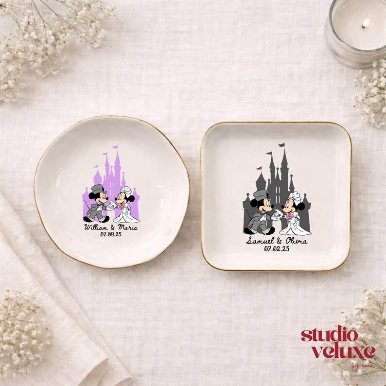 May include: Two white ceramic ring dishes with gold rims. One is round, the other square. Each features a cartoon couple in wedding attire in front of a castle. The round dish says "William & Maria 07.09.25". The square dish says "Samuel & Olivia 07.02.25".
