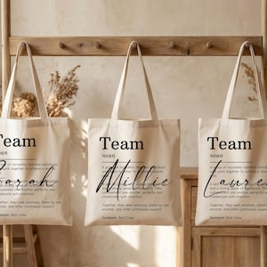 Peut inclure: Trois sacs fourre-tout en toile beige avec le mot "Team" imprimé en noir. Chaque sac porte un nom différent en écriture cursive : "Sarah", "Millie" et "Lauren". Les sacs ont également une définition de "Team" imprimée en noir.