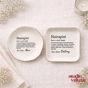 May include: Two white ceramic trinket dishes with gold-toned rims. One is circular, the other square. Both feature black printed text defining the noun Hairapist as a scissor wielding, miracle working genius. The circular dish lists Alexa, while the square dish lists Britney as a see also reference.