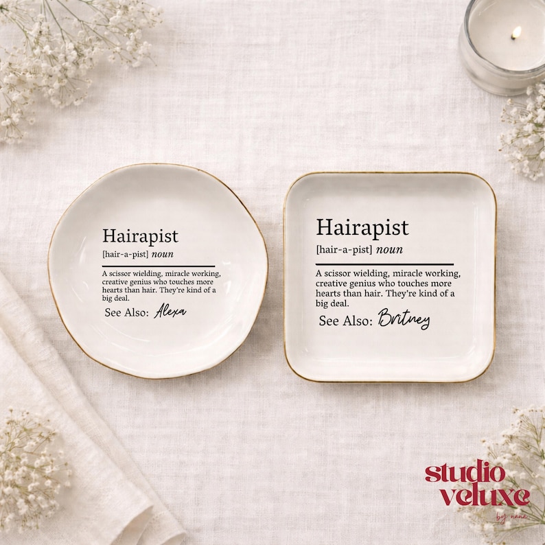 May include: Two white ceramic trinket dishes with gold-toned rims. One is circular, the other square. Both feature black printed text defining the noun Hairapist as a scissor wielding, miracle working genius. The circular dish lists Alexa, while the square dish lists Britney as a see also reference.