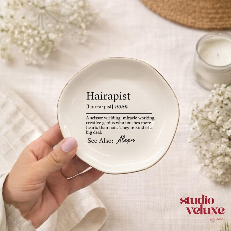 May include: A round white ceramic trinket dish with a gold-toned rim. The center features black text that reads: Hairapist [hair-a-pist] noun. A scissor wielding, miracle working, creative genius who touches more hearts than hair. They're kind of a big deal. See Also: Alexa.