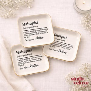 May include: Three square white ceramic trinket dishes with gold-toned rims. Each dish features black printed text defining the word Hairapist as a scissor wielding, miracle working, creative genius. Each dish includes a unique name: Millie, Britney, or Kaitlyn.