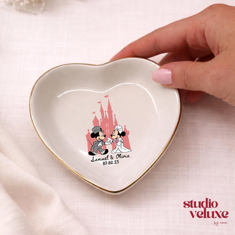 May include: Heart-shaped trinket dish with a gold rim. The white ceramic dish features a pink castle illustration and a cartoon couple, with the names "Samuel & Olivia" and the date "07.02.25" printed below. The dish is held by a hand.