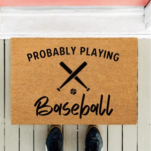 May include: A rectangular doormat made of natural-colored coir fiber. The mat features the words "PROBABLY PLAYING" above a crossed baseball bat and a baseball graphic, with the word "Baseball" below. Black text on a brown background.