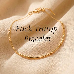 May include: Gold-coloured bracelet with the text "Fuck Trump Bracelet" in black. The bracelet features a delicate chain with small, cylindrical and spherical beads. The background is a beige fabric.