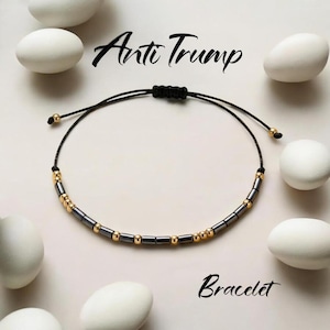 May include: Black cord bracelet with gold and grey beads. The text "Anti Trump" is written above the bracelet, and the word "Bracelet" is written below it.