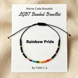 May include: A Morse code bracelet on a white display card labeled Morse Code Bracelet, LGBT Beaded Bracllet, Rainbow Pride, and By YOMI C.S. The bracelet features black cord, small black beads, gold-tone spacers, and a sequence of rainbow-colored beads.