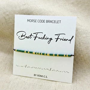 May include: A Morse code bracelet with green and gold beads on a black cord, displayed on a white card. The card reads "MORSE CODE BRACELET" and "Best Fucking Friend" in cursive. The bottom of the card says "BY YOMI C.S."