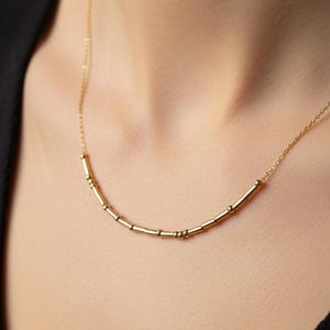 May include: A delicate gold necklace with a series of small, cylindrical beads and accents. The necklace is worn against a black top, highlighting the jewelry's warm, metallic color. The design is simple and elegant.